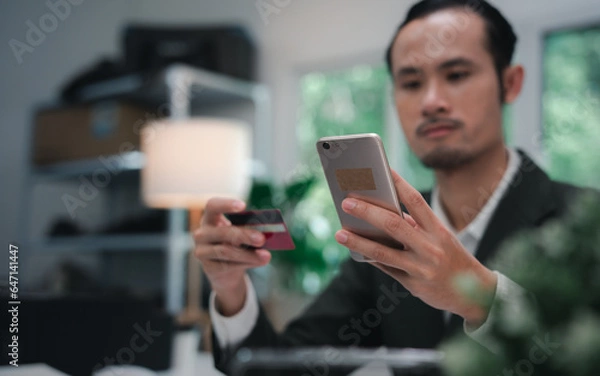 Fototapeta Asian businessman smiling and happy doing online shopping outside office building, man using mobile phone, holding bank credit card