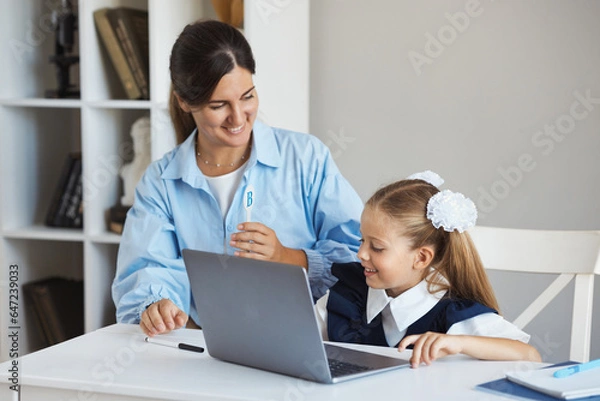 Fototapeta Tutor teaches little schoolgirl 7-8 years old English and gives exams on laptop, sitting at a table in a classroom during lesson. A teacher helps and explains a task to student on a computer at school