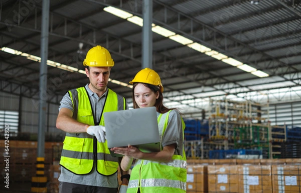 Fototapeta Female engineer in helmet and safety vest holding latop discussing with male engineer in factory.