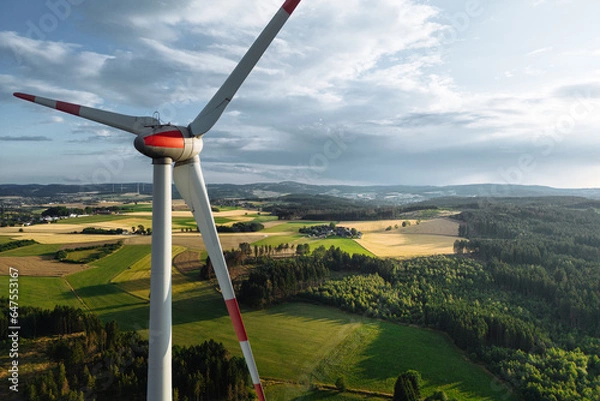 Fototapeta Wind turbines in a hilly forest in front of a partly cloudy, but sunny sky are seen from an aerial view during sunset