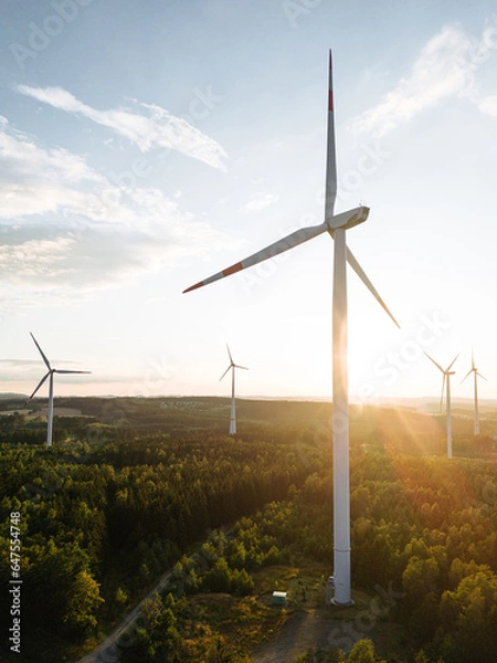 Fototapeta Wind turbines in a hilly forest in front of a partly cloudy, but sunny sky are seen from an aerial view during sunset