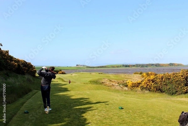 Fototapeta A young man swinging a golf club on a tee box surrounded by the beautiful views of the sea in the background, outside Inverness, Scotland