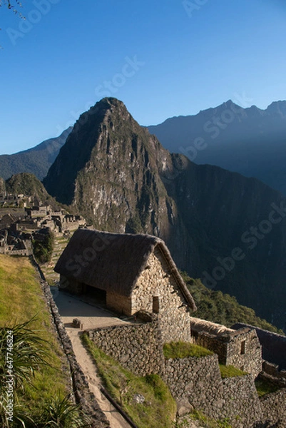 Fototapeta Entrance to Machupicchu, view of a stone house surrounded by the archaeological site with mountains and a blue sky and sunbeam, Sacred Valley, Peru. 