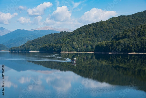 Fototapeta Small boat glides across the surface of lake surrounded by green forest hills and amazing reflection of cloudy sky. Vidrarudam. Trasfagarasan road. Romania
