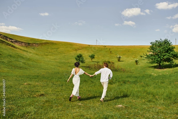 Fototapeta young couple of newlyweds running in green field, bride and groom in wedding gown holding hands