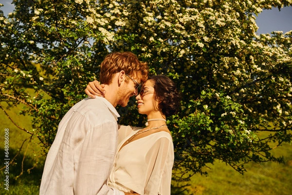 Obraz newlyweds couple in countryside, asian bride in white dress and groom hugging near big tree