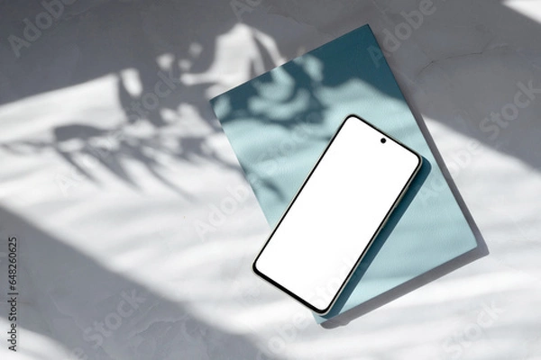 Fototapeta Mobile telephone empty display mock up, blue notebook on white marble table with natural abstract sun light shadows background. Aesthetic business brand template
