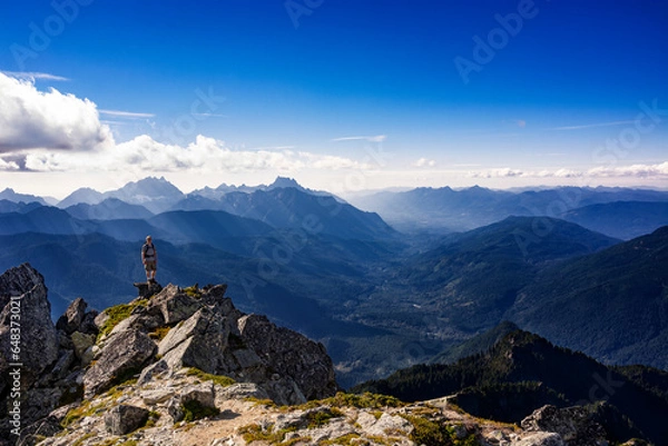 Obraz Adventurous athletic male hiker standing on top of a rugged mountain in the Pacific Northwest with jagged mountains in the background.
