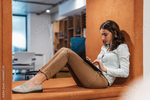 Fototapeta Girl sitting at recessed wall area working on her lap top at modern office room.
