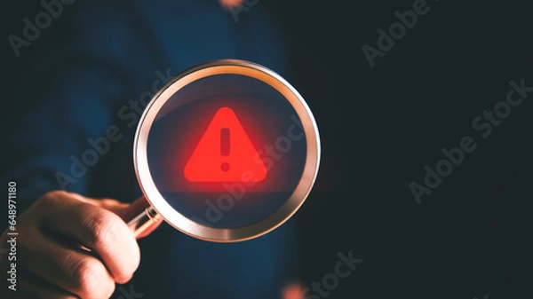 Fototapeta Red warning icon on a magnifying glass lens held by the businessman hand.