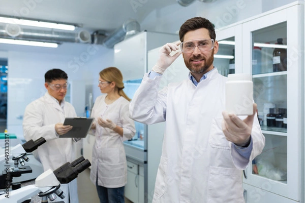 Fototapeta Portrait of a smiling young man pharmacist standing in a laboratory in a uniform and holding a plastic jar with material in his hands. Shows the result to the camera. Colleagues are discussing behind.
