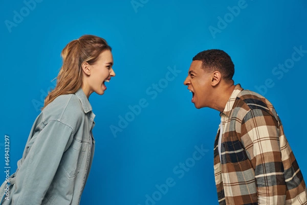 Fototapeta multicultural couple screaming at each other on blue backdrop, arguing and misunderstanding