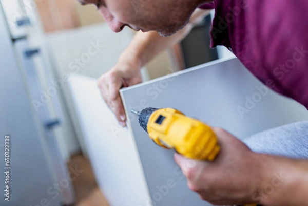 Fototapeta Man holding electrical screwdriver while assembling kitchen wooden elements.