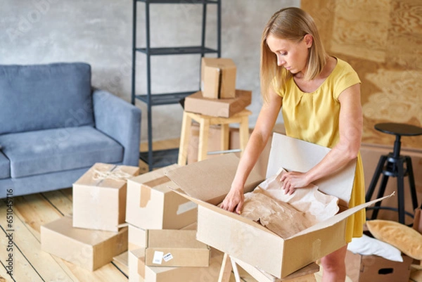 Fototapeta Young girl packing plates into the boxes ready to move. Woman unpacking moving boxes in her new home. unpack personal stuff from carton boxes.