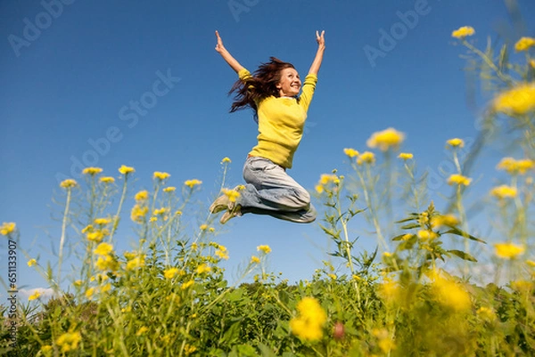 Obraz Happy and beautiful young woman in a bright yellow sweater and blue jeans  jumping high in a sunny summer field.