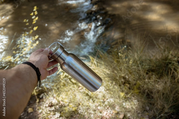 Obraz Hand taking an aluminum thermal bottle to hydrate on a hiking trail