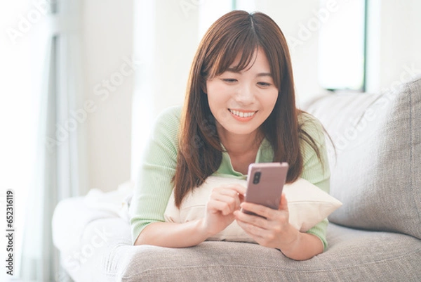 Obraz リビングのソファに横になってスマートフォンを操作しながらくつろぐ女性