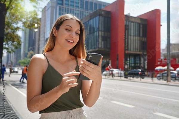 Fototapeta Portrait of beautiful Brazilian girl using mobile phone app for chat on sunny day on Paulista Avenue in Sao Paulo, Brazil