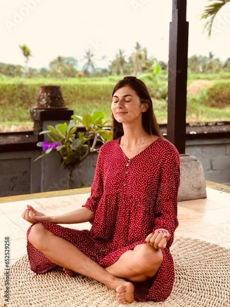 Fototapeta Young beautiful woman meditating and doing yoga in red dress in abandoned temple with green palms and rise field background on Bali. Female practicing pose. High quality FullHD vertical footage
