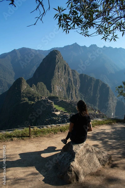 Fototapeta Young woman with long flowing hair, wearing sportswear, sitting on a rock watching the archeological site of Machupicchu, Peru. Views of the mountains, with a clear and sunny sky. 