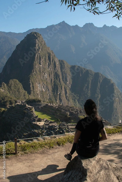 Fototapeta Young woman with long flowing hair, wearing sportswear, sitting on a rock watching the archeological site of Machupicchu, Peru. Views of the mountains, with a clear and sunny sky. 