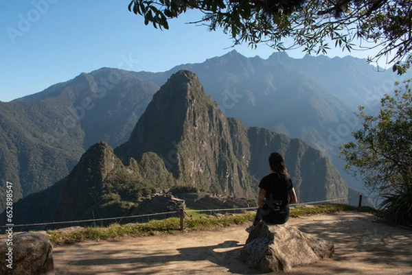 Fototapeta Young woman with long flowing hair, wearing sportswear, sitting on a rock watching the archeological site of Machupicchu, Peru. Views of the mountains, with a clear and sunny sky. 