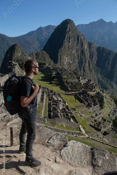 Fototapeta Male backpacker standing with backpack, reusable water bottle, long hair pulled back, observing the archeological site of Machupicchu, Peru. 