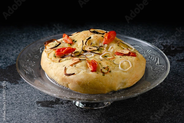 Fototapeta Focaccia with onion, cherry tomatoes, garlic and spices. Close-up, glass plate, on marble, granite table, black backgroun