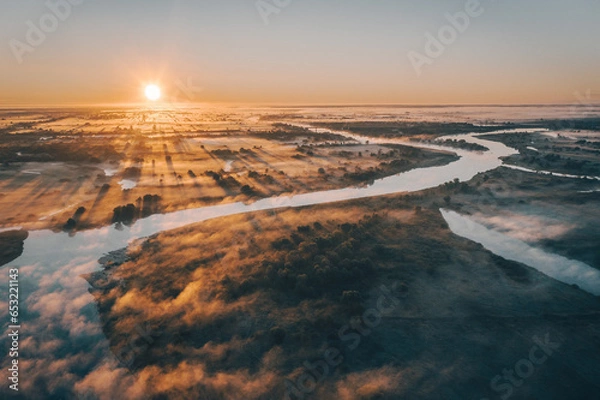 Fototapeta Morning sunrise on the big river. The photo was taken from a drone, there is beautiful fog and a blue winding river below.  View of a field next to a river.