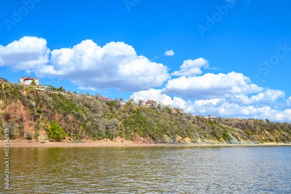 Fototapeta River embankment with buidlings on a fine spring day. Distant view from the river. Blue cloudy sky and calm river water