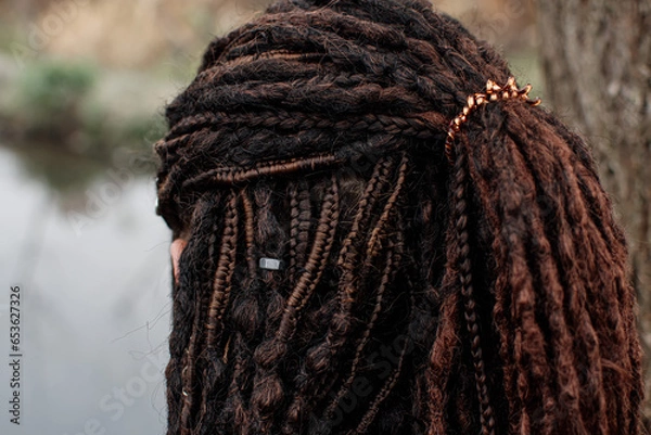 Fototapeta A girl with dark hair stands inquiringly at the camera. A walk in nature. The girl has many thin braids on her head