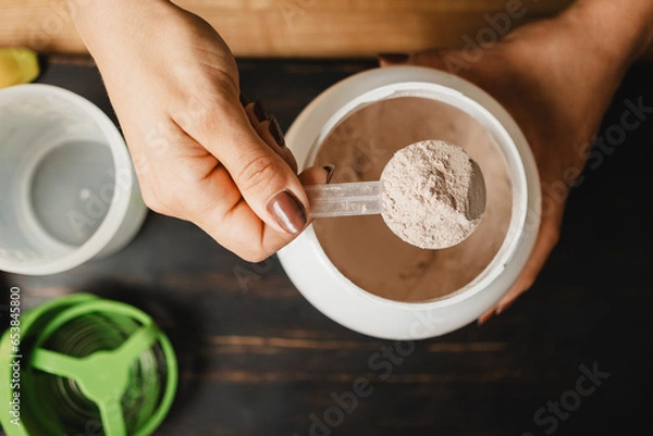Obraz Female hand holds measuring spoon with portion of whey protein powder above plastic jar on wooden table with shaker, banana and apple fruit. Process of making protein drink, top view