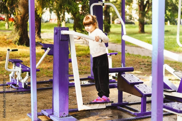 Obraz Happy little girl training on a simulator on an outdoor sports ground. A smiling, cute five-year-old girl doing exercises on a pendulum exercise machine outdoors in the park. Healthy lifestyle concept