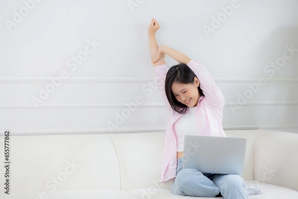 Obraz Beautiful young asian businesswoman sitting on sofa and working from home and break while stretch arm for relax in living room at home, woman is freelance working on laptop computer, business concept.