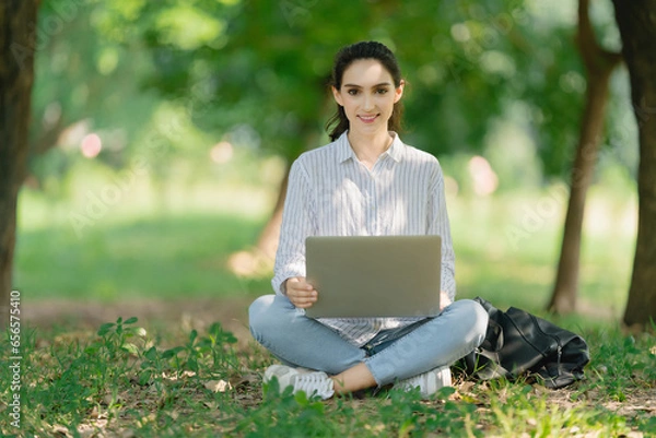 Fototapeta Smiling young Woman laughing at her laptop computer in the park. Women studying in an outdoor, working on computer or video meeting.