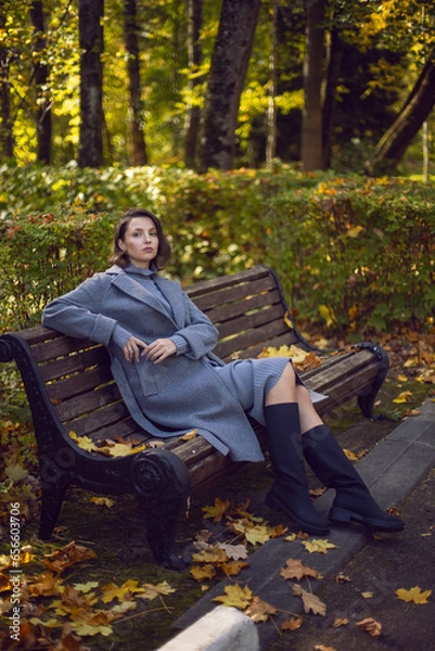Obraz brunette woman in a gray coat and boots is sitting on a park bench in autumn among the trees. Orange foliage and a wooden bench during the day
