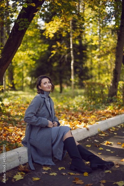 Obraz brunette woman in a gray coat and boots is sitting on a park curb in autumn among the trees. Orange foliage and a wooden bench during the day