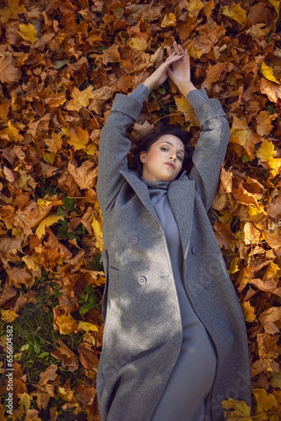 Obraz brunette woman in a gray coat and boots it lies on the foliage in autumn. Orange foliage during the day