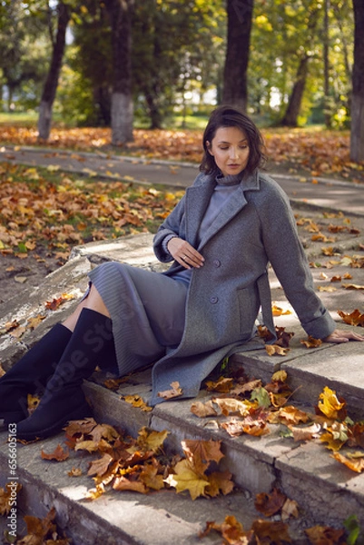 Obraz brunette woman in a gray coat and boots is sitting on the stone steps in autumn among the trees. Orange foliage during the day