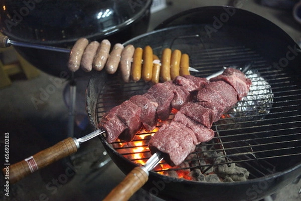 Obraz 炭火の上で香ばしく焼き上げ、肉汁が滴るジューシーな質感に仕上げたバーベキューの串焼き肉。しっとりした赤身の輝きが際立つ一景で、アウトドアの醍醐味や宴会、夏のレジャー向けの販促に最適な高品質素材です。 | Juicy beef skewers and sausages being grilled over a charcoal BBQ fire.
