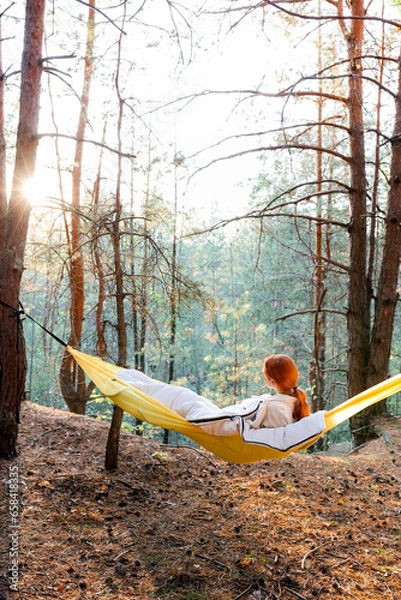 Fototapeta A girl in a hammock in an autumn pine forest looks at the sunset