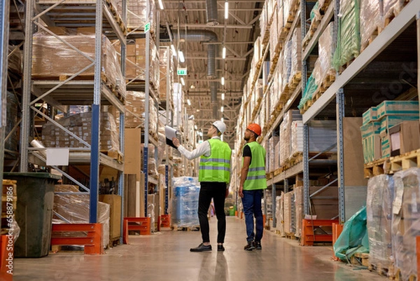 Fototapeta two men in warehouse wearing safety helmet, vest. Concept for industry, job, meeting, work training. Two caucasian warehouse workers walking in distribution storage area discussing