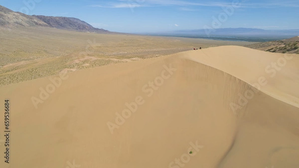 Fototapeta A large sand dune in the middle of the steppe. Drone view of a huge pile of sand. Tourists are walking, enjoying the view. A river runs in the distance and grass grows. The sky with white clouds