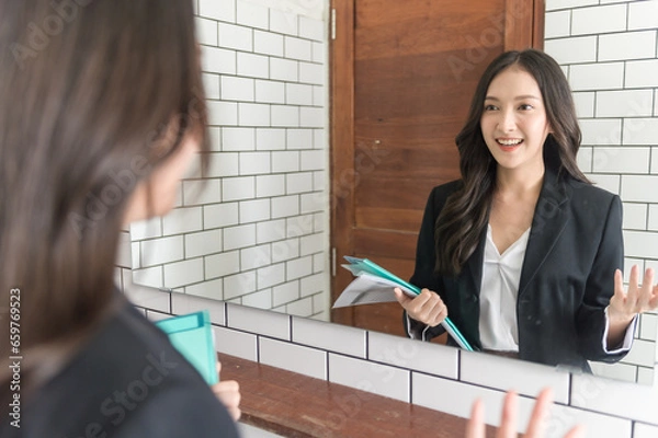 Fototapeta Young beautiful Asian woman in formal suit checking dressing tidiness and encourage herself in mirror at home before go for job interview. Woman recruitment employee or staff in company concept.