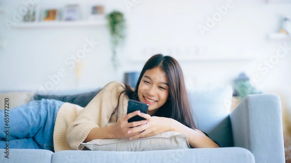 Obraz Happy young asian woman relax on comfortable couch at home texting messaging on smartphone, smiling girl use cellphone, browse wireless internet on gadget, shopping online from home