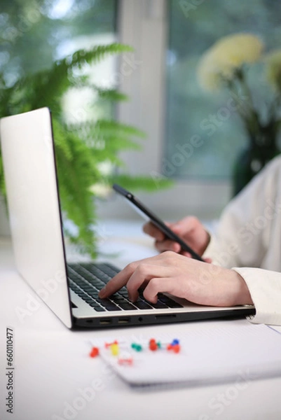 Fototapeta Online shopping concept. There is a white laptop on a white table. next to it is a notepad for notes and a worker’s hands on a keyboard and smartphone
