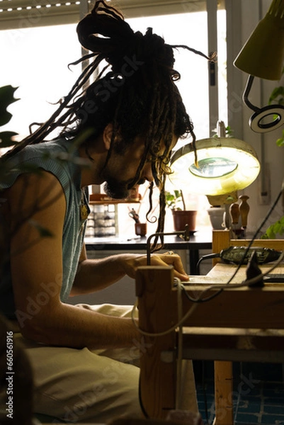 Obraz Young Rastafarian artisan jeweler working in his home workshop. Vertical photo. Concentrated at work.