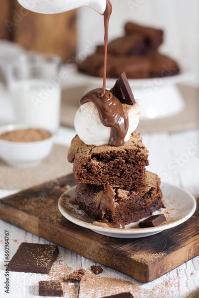 Fototapeta Brownie with a scoop of ice cream cut into pieces and topped with chocolate from a gravy boat. In the background is a northerly table with milk and the rest of the brownies.