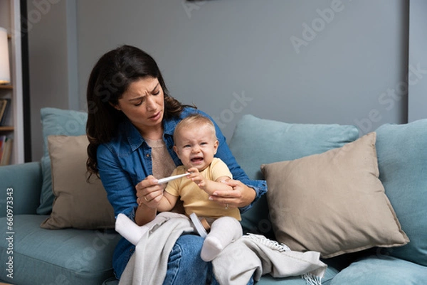 Fototapeta Mother measuring temperature of her sick crying baby sitting on sofa in living room, little infant child catching cold, having influenza or grippe. Young mom worry for her kid cry