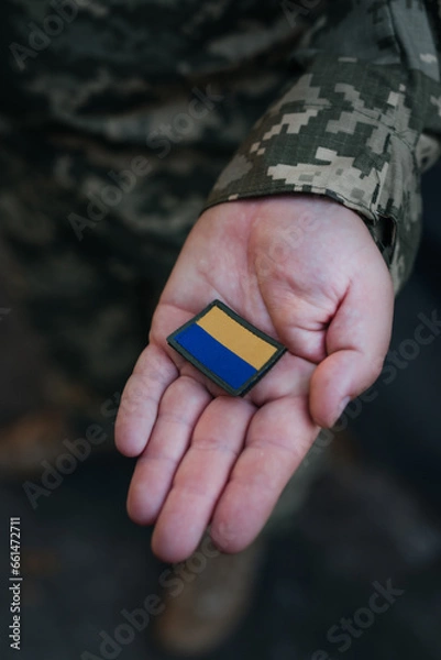 Fototapeta A Ukrainian military man holds the emblem of the national flag in his hand as a symbol of strength. War in Ukraine. Dramatic background, warm colors